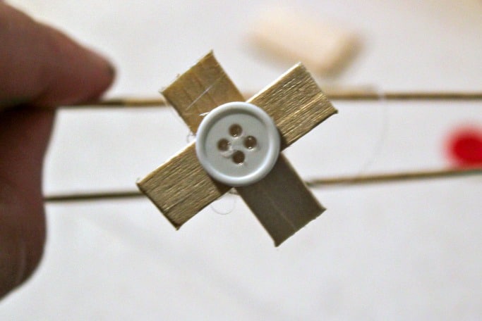 步骤4：将冰棒棍的中间长片切成两半。粘在一起成X形，作为飞机的螺旋桨。 在螺旋桨中心贴上一颗纽扣。