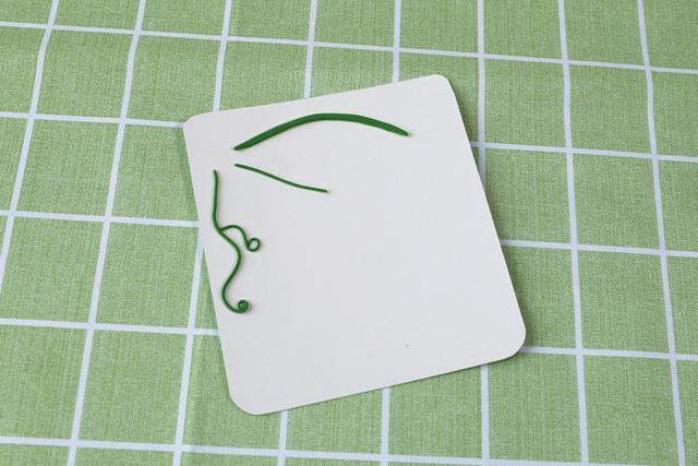 步骤2：用绿色粘土揉出粗细不等的长条，在卡纸一角做好造型粘在上面