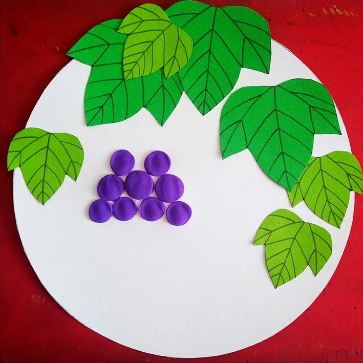 步骤3：按照自己预先设想的构图把叶子粘贴到圆形卡纸上，并在合适的位置用紫色橡皮泥粘贴出葡萄。