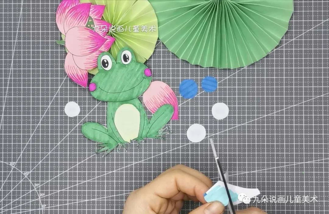 步骤5：将小青蛙和荷花沿着轮廓线剪下，再用剩余的白纸剪一些大小不同的圆形，有的可以涂成蓝色，有的涂黄色，有的是白色，来表示荷叶上的圆滚滚的露珠。